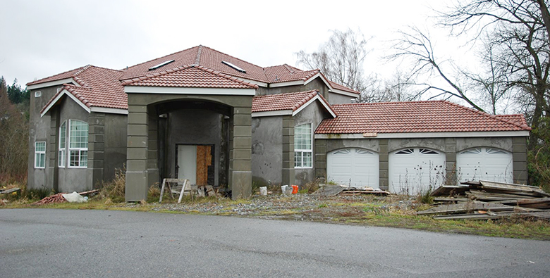 An image of a new home construction that appears to have most of the exterior completed but which clearly is not finished and has been abandoned for some time.