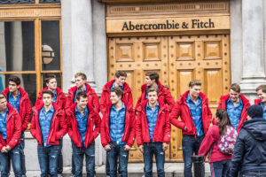 Two lines of 13 male models, wearing the same outfit: jeans, a blue button down shirt, and a red sweater. They are standing in front of the doors of an Abercrombie and Fitch store.