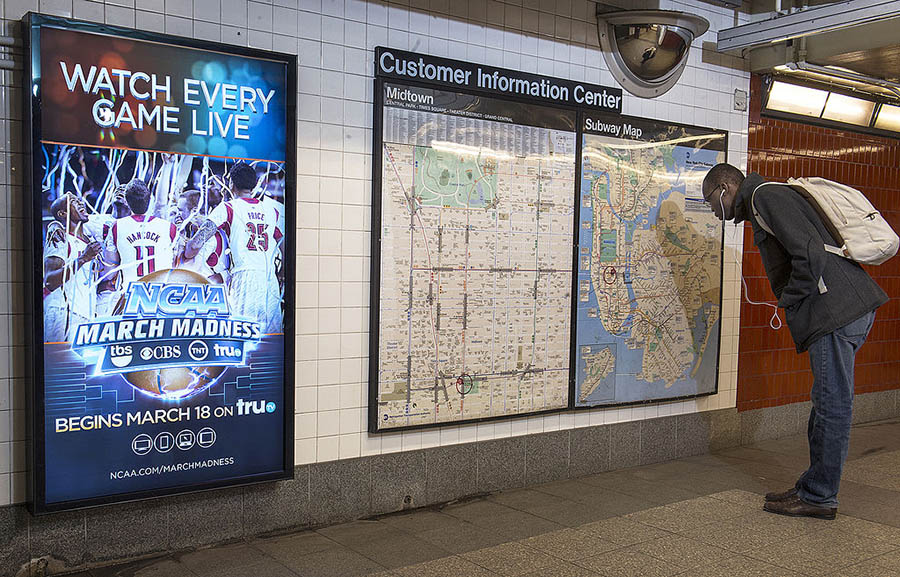 A photograph of a wall in a subway in New York. The left side of the wall features a vertical, rectangular television screen showing an advertisement for NCAA March Madness. The right side of the wall features a large bulletin board labeled “Customer Information Center,” showing two different maps. A person with a backpack is to the right of the bulletin board, bent over reading the map.