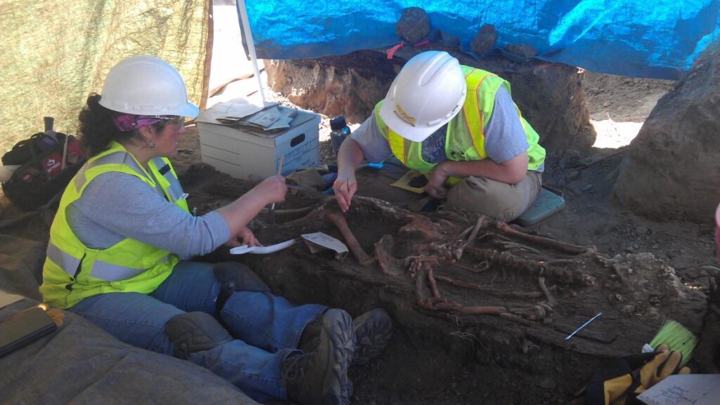 Two archaeologists are seated in the dirt, excavating a human skeleton under a blue tarp.