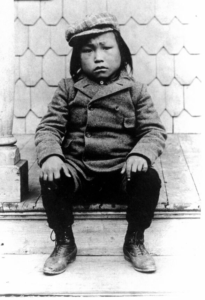 A black and white photo of a small boy sitting on the steps of a building.