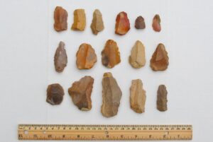 Three rows of small chipped stone tools of various colors and shapes are arranged above a ruler.