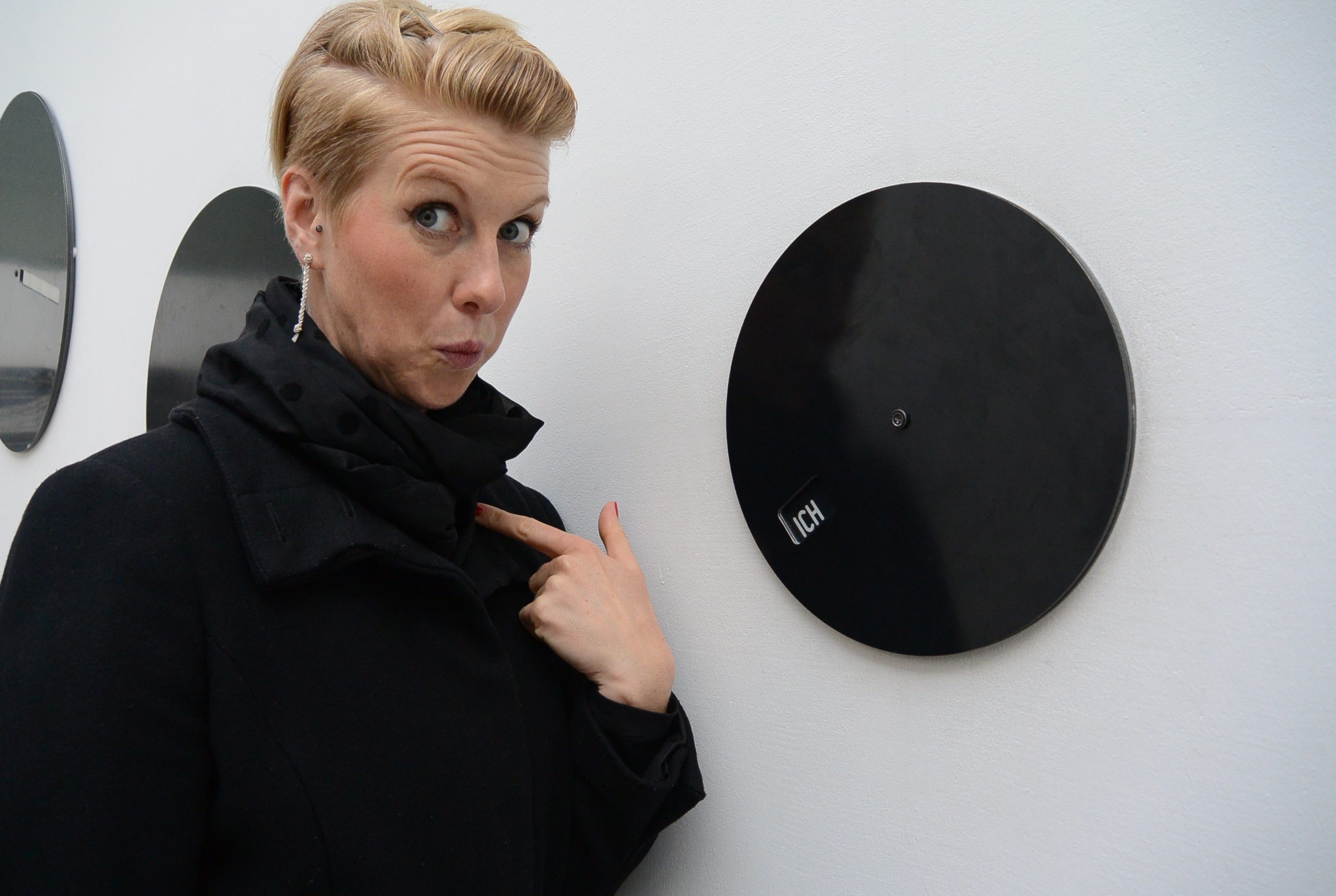 A feminine presenting person pointing to themself next to a record disk mounted on a wall.