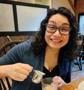 This is a photo of Sajonna Sletten. She is smiling as she pours cream into a coffee mug. The background is a cafe.